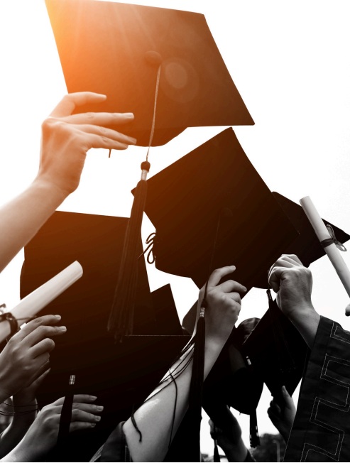 Graduates raising black mortarboards and holding diplomas against a bright sky with sunlight flare.