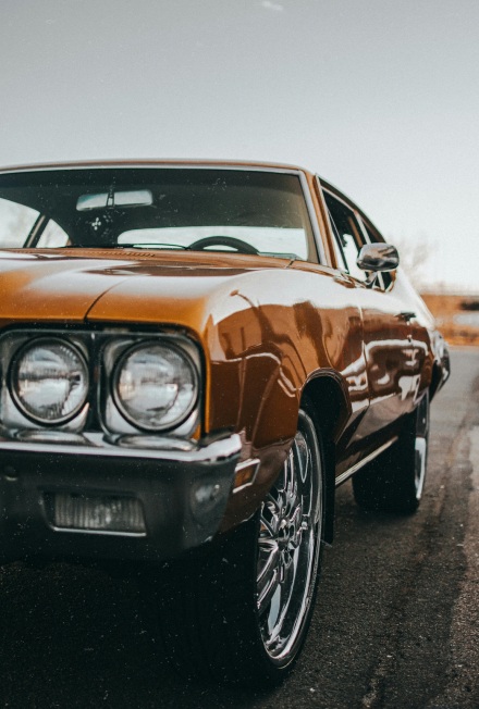 Close-up of the front side of a vintage brown car with chrome rims parked on a street.